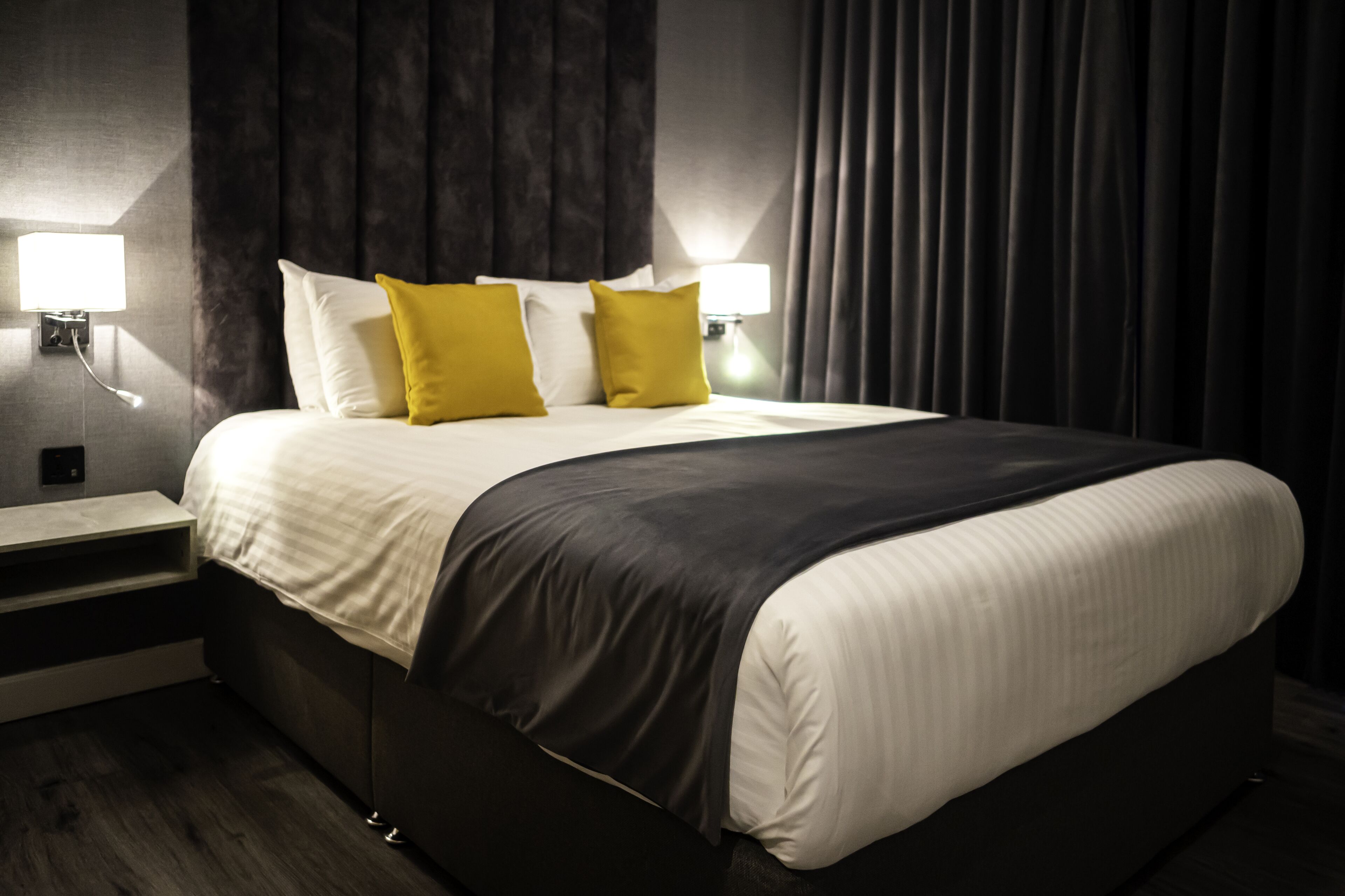 A modern hotel room with a large bed, two bedside lamps, and a dark curtain backdrop.