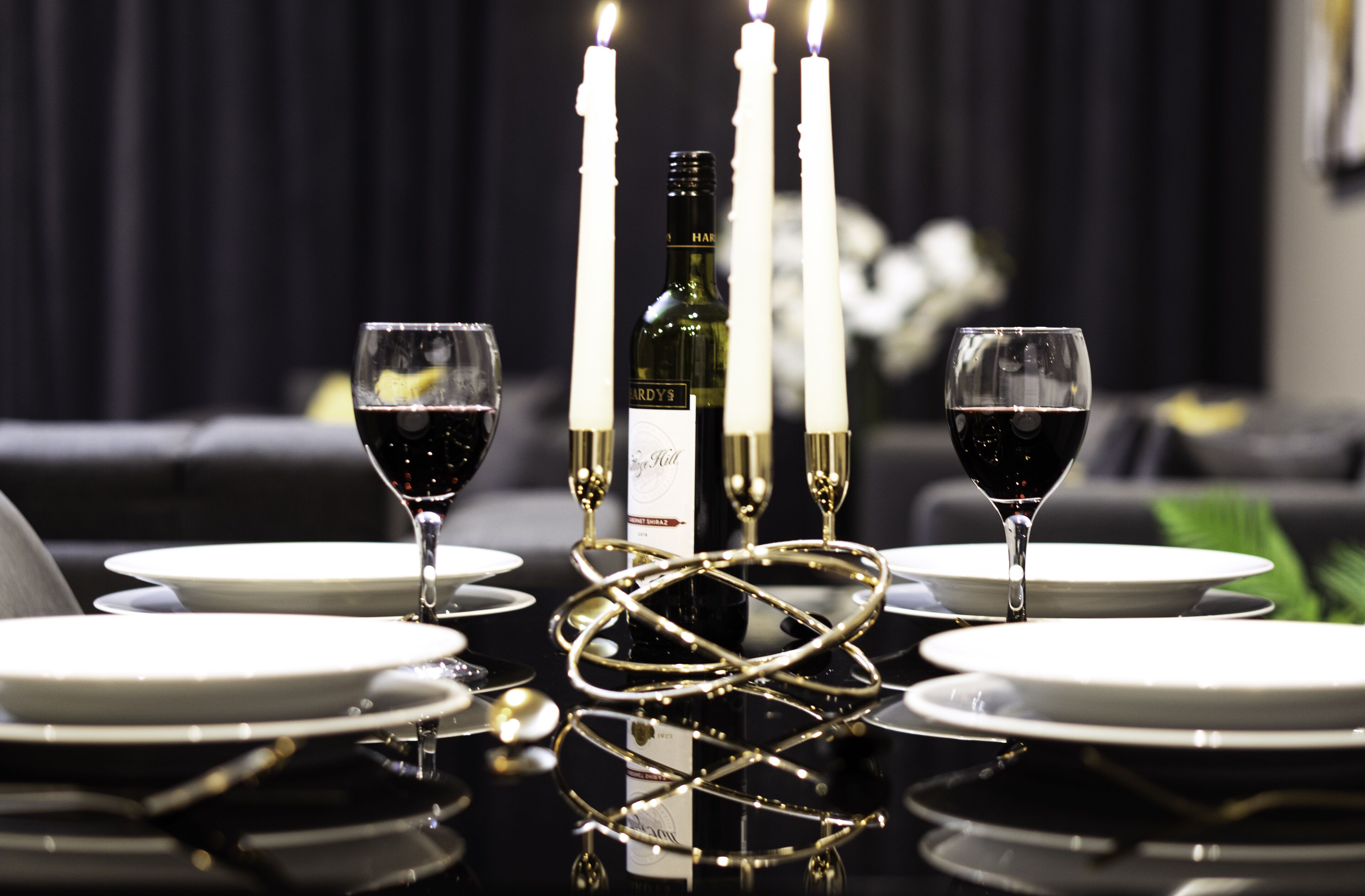 A dining table set with white plates, two wine glasses, a bottle of wine, and four lit candles.