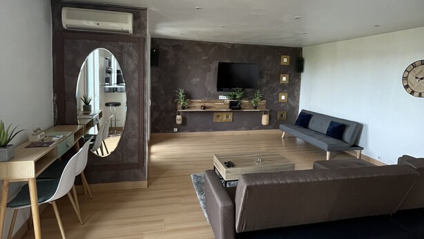 A modern living room with a wooden floor, a sofa, a dining area with a table and chairs, a TV, and a wall-mounted clock.