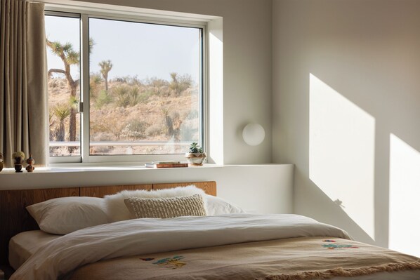 A bedroom with a large window, a bed with white bedding, a wooden headboard, and a small plant on the windowsill. - Panorama House: Featured in Dwell (Yucca Valley)