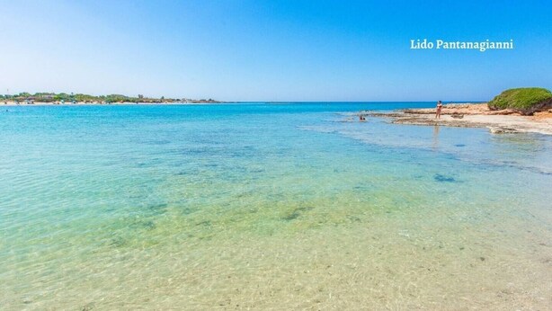 Une plage dégagée avec une eau peu profonde, une personne debout sur le rivage et une ligne de côte lointaine avec des bâtiments.