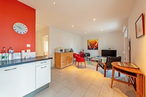 Une cuisine moderne avec des meubles blancs, un mur d’accent rouge et une horloge. On y trouve également un coin repas avec une table, des chaises et un téléviseur.