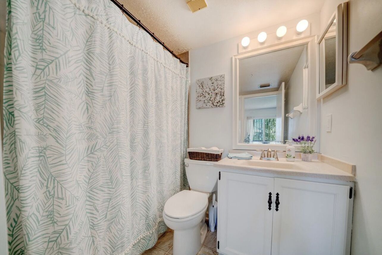 A bathroom with a white toilet, a white cabinet with black handles, a white countertop with a mirror, and a shower curtain with a leaf pattern.