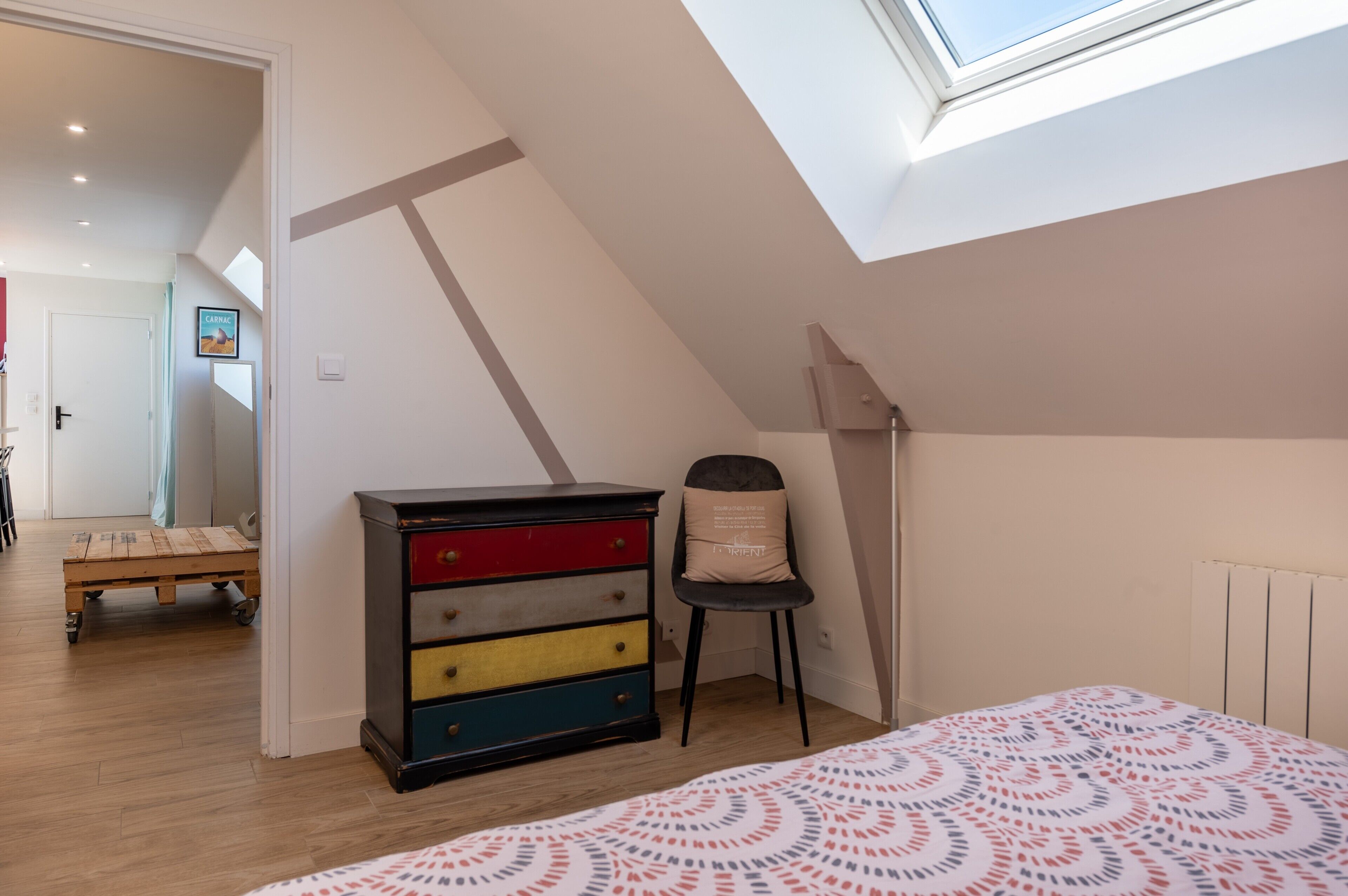 A bedroom with a sloped ceiling, a bed with a patterned bedspread, a dresser, a chair, and a small table.
