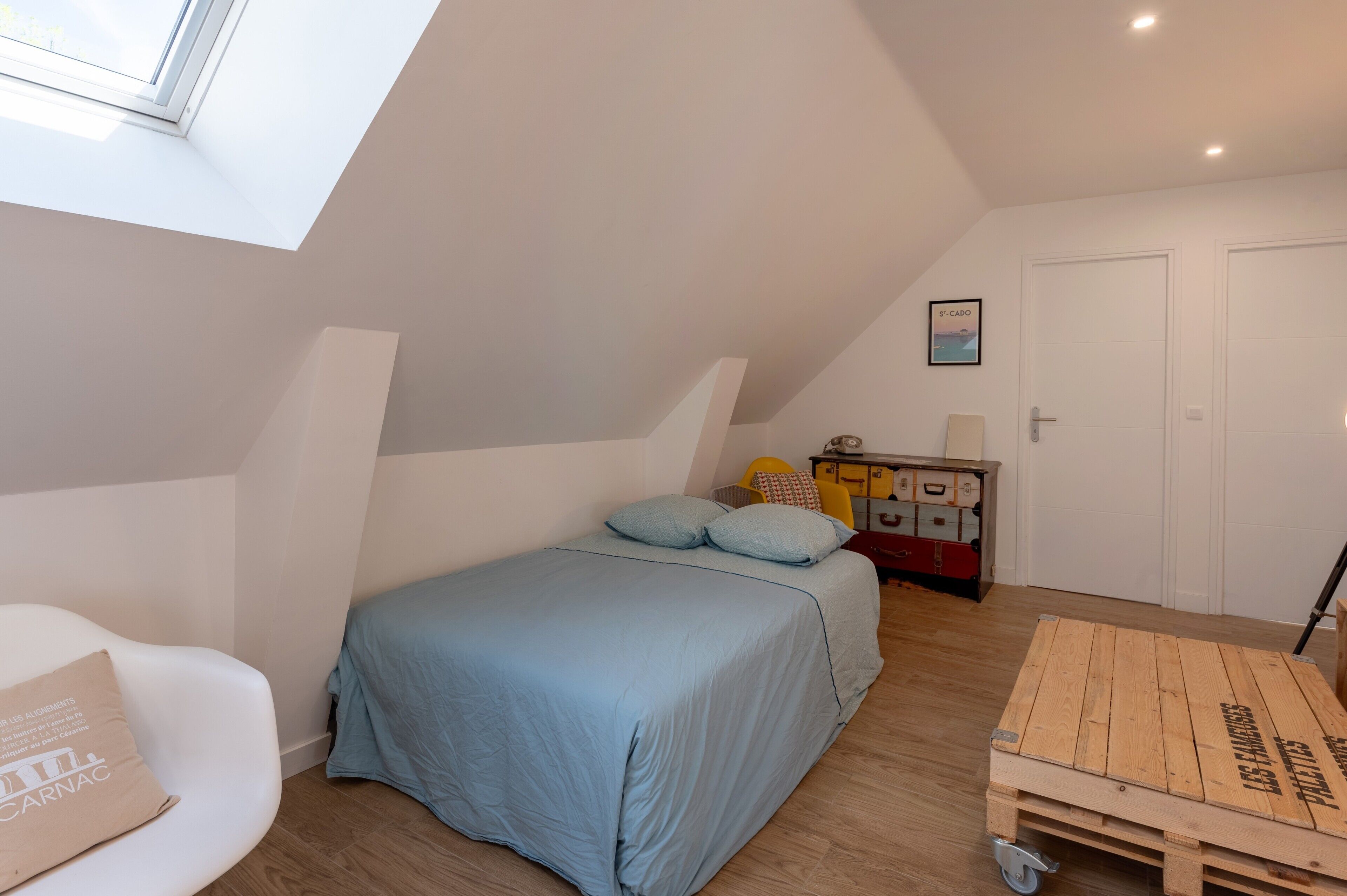 Una camera da letto con soffitto spiovente, un letto con lenzuola blu, una sedia bianca, un tavolino e una cassa di legno.