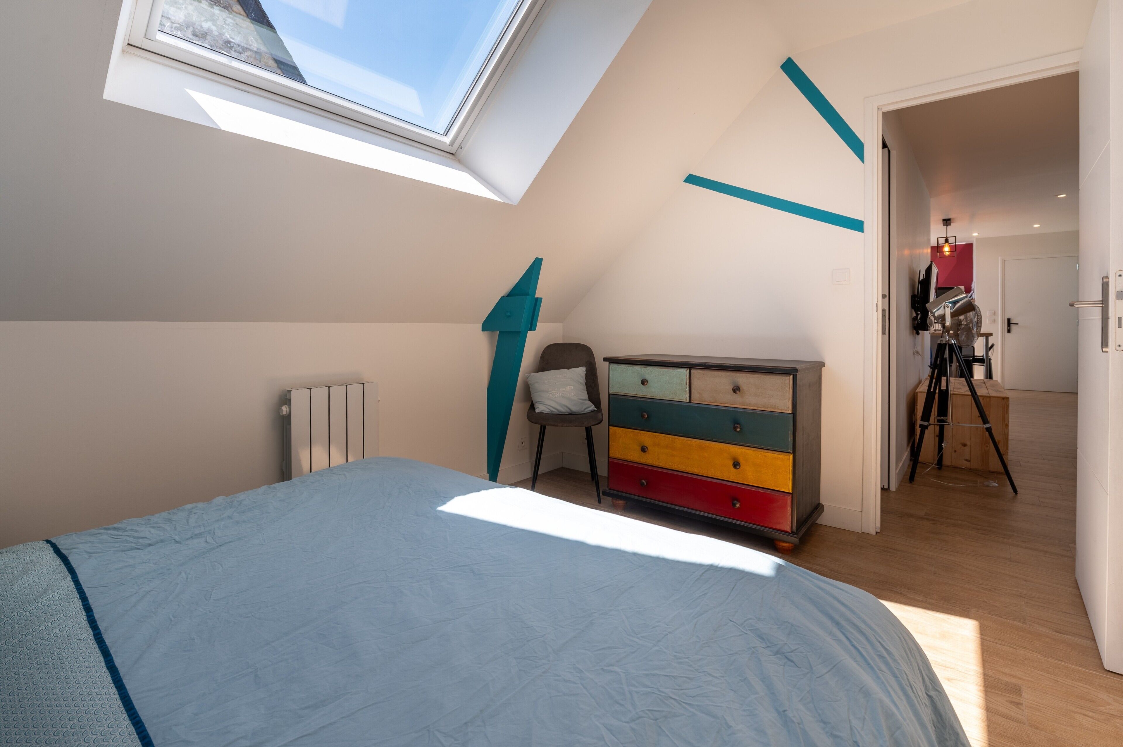 Une chambre avec un puits de lumière, une commode aux couleurs vives et un trépied en arrière-plan.