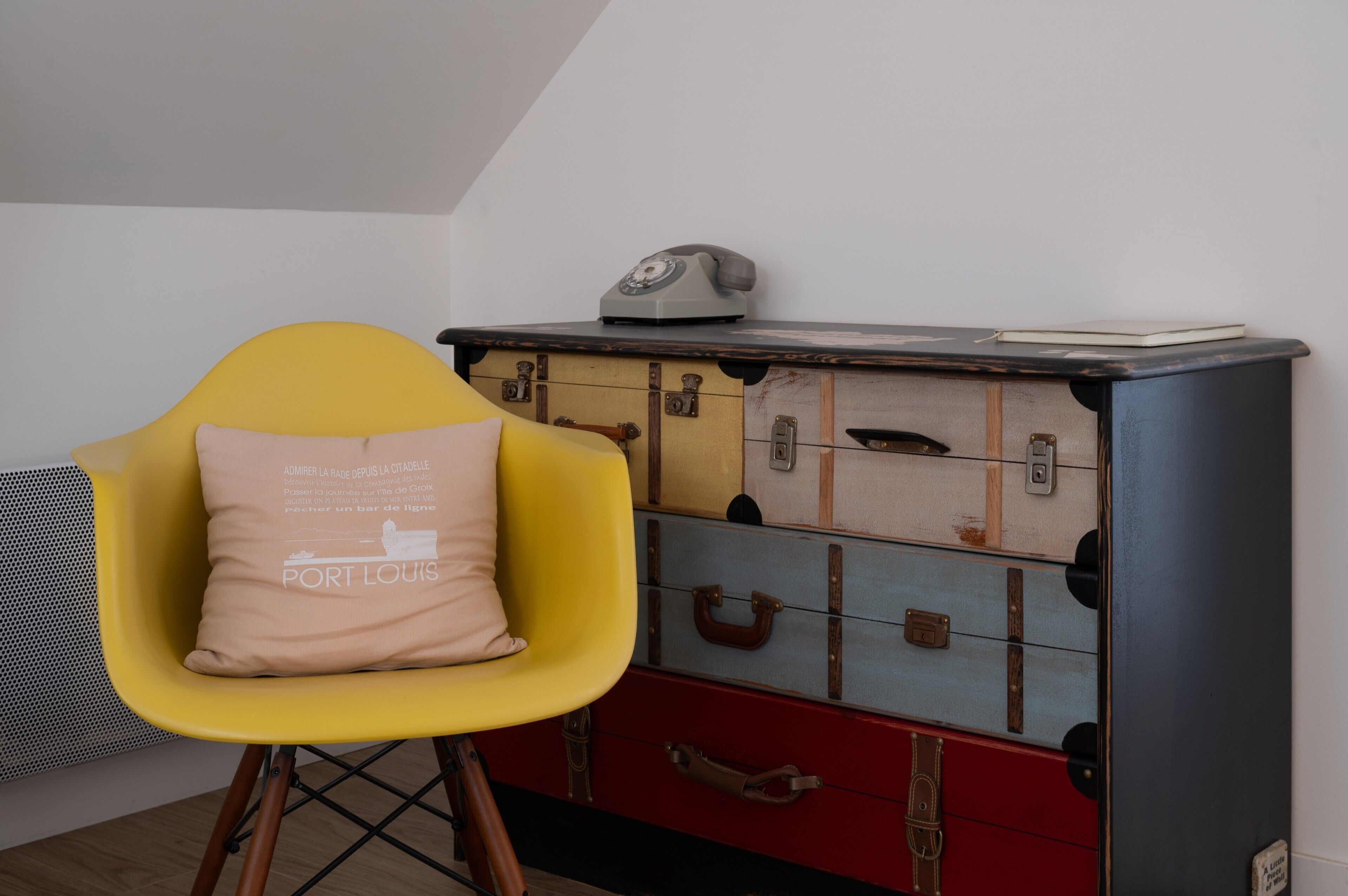 A room with a yellow chair, a chest of drawers with multiple compartments, and a vintage telephone on top.