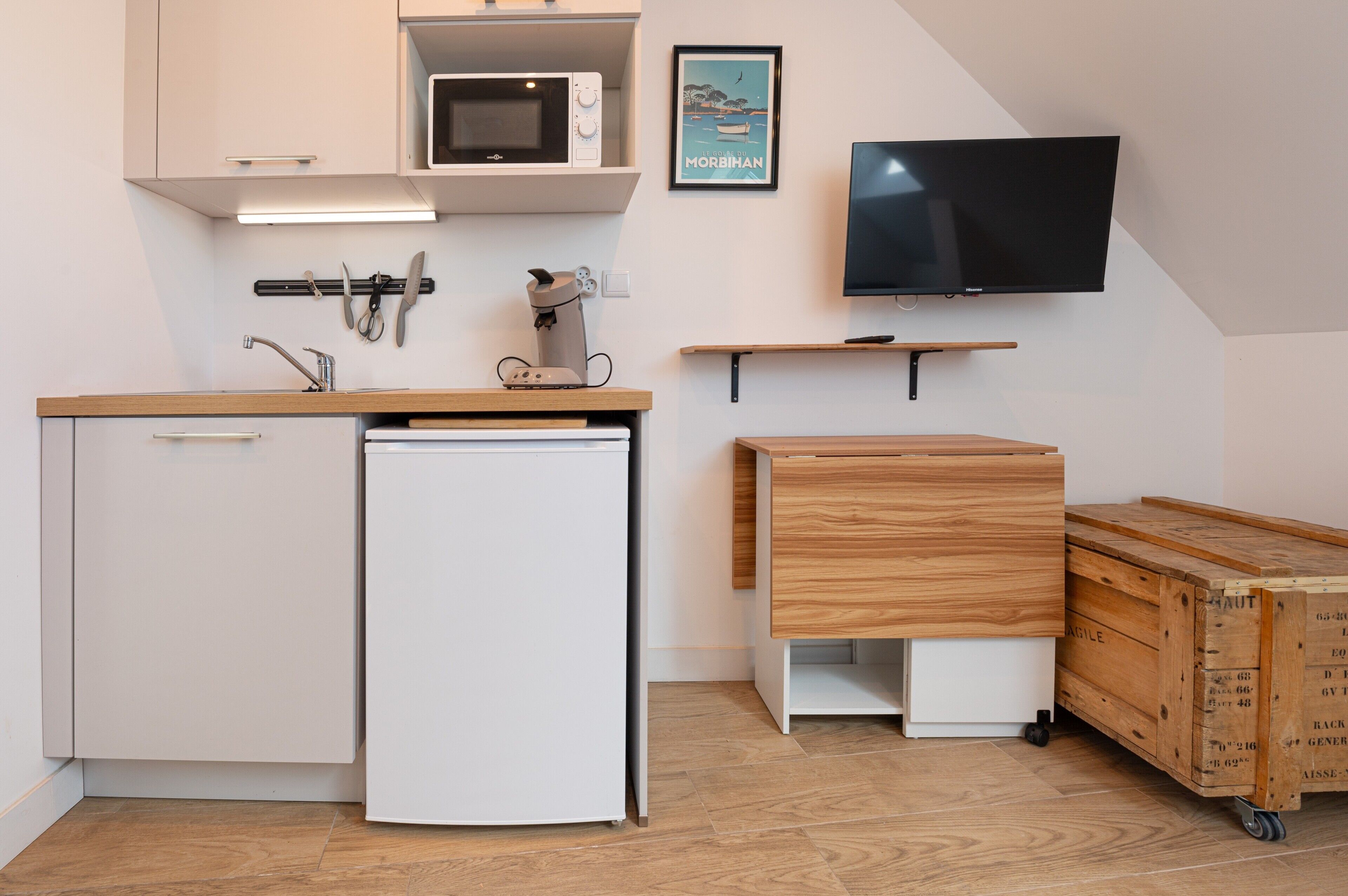 A compact kitchen with a microwave, a small refrigerator, a wooden cabinet, a television, and a wooden chest of drawers.