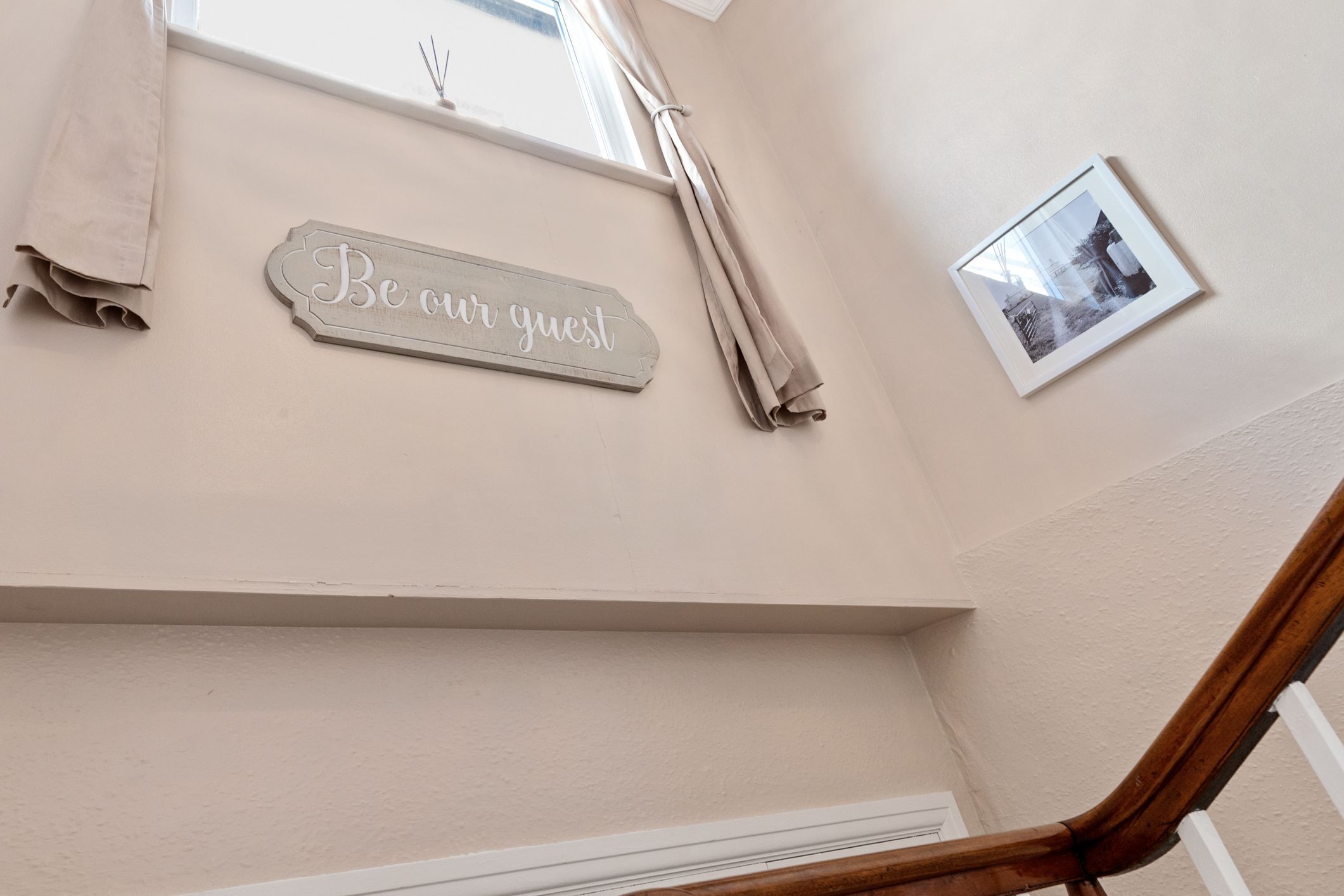 Una pared con un letrero que dice 'Be our guest', una foto enmarcada y una ventana con cortinas.