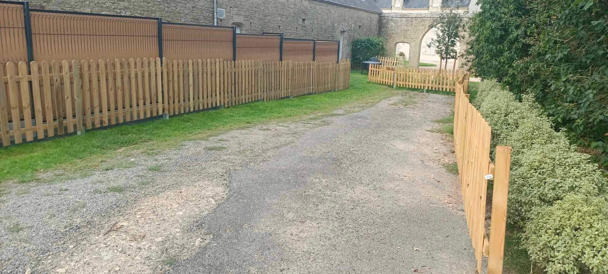 Een grindpad leidt naar een houten hek, een stenen gebouw en een groene haag.