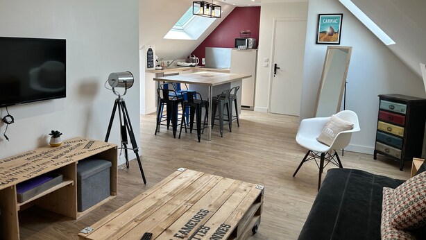 Un espacio habitable pequeño con televisión, una mesita de madera, zona de comedor con sillas, sofá y kitchenette.