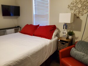 Quarto com cama, televisão na parede, mesa de cabeceira com abajur e uma cadeira.