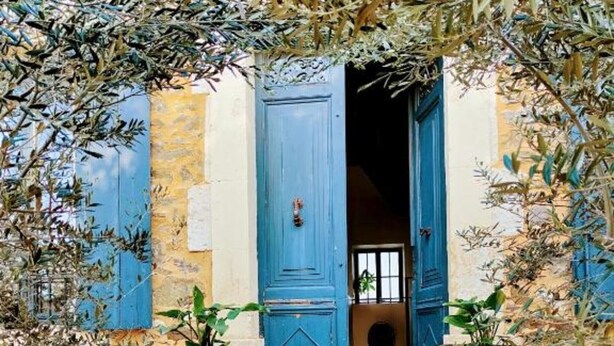 Un escalier en pierre menant à une porte bleue, de part et d’autre de laquelle se trouvent des plantes en pot et des branches d’olivier.