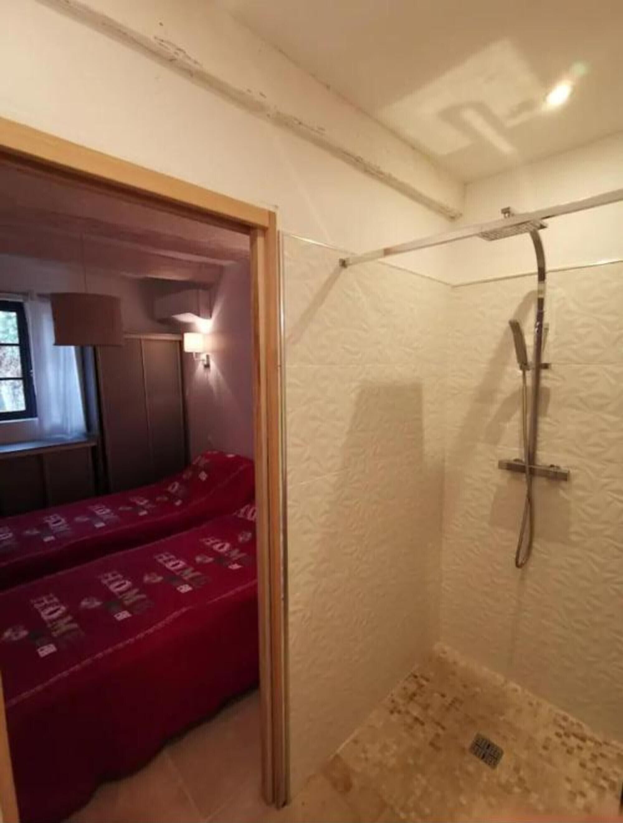 Una habitación pequeña y funcional con ducha, una cama con manta roja y una ventana con cortinas.