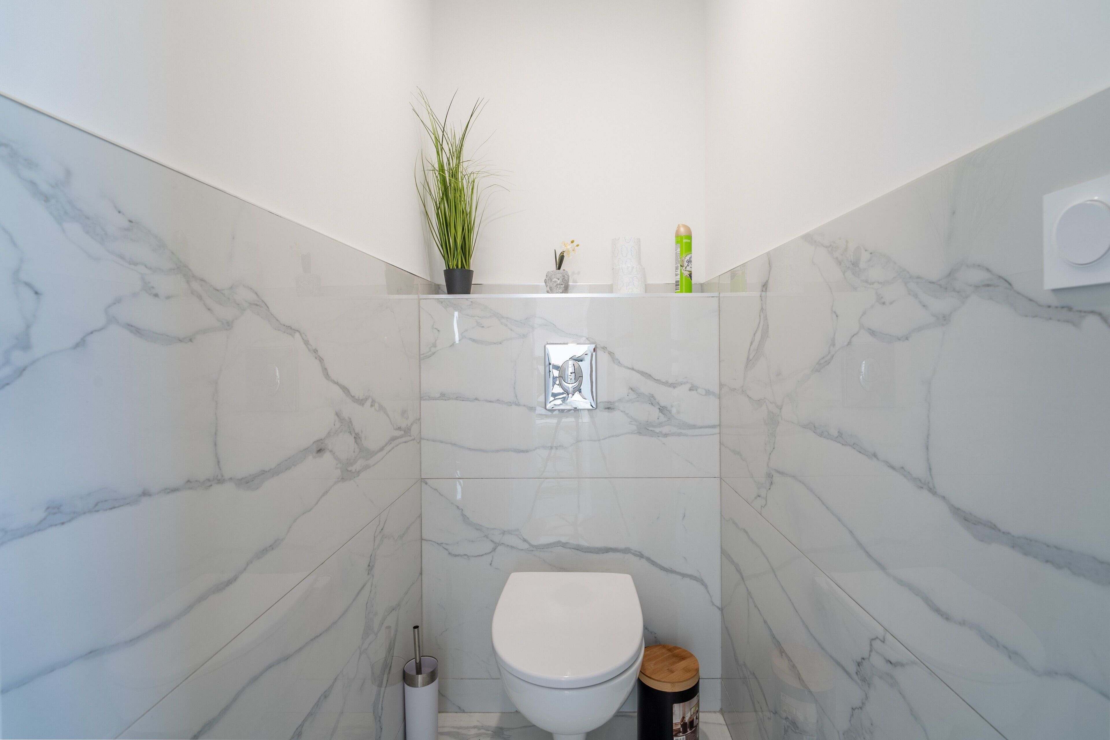 Un baño con inodoro blanco, pared de mármol, planta en maceta y estante con artículos de aseo.