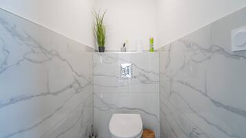 Un baño con inodoro blanco, pared de mármol, planta en maceta y estante con artículos de aseo.
