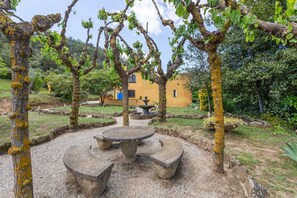 Un patio con una mesa de piedra y bancos, rodeado de árboles y un edificio amarillo al fondo.