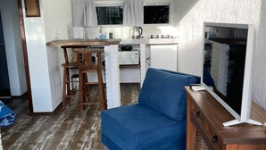 A compact living space with a kitchenette, a dining area, and a seating area with a blue sofa and a wooden desk with a computer monitor. - Altos da Lagoa-praia do Rosa,ibiraquera Studio 1 Para Casais Calmo e Tranquilo (imbituba)