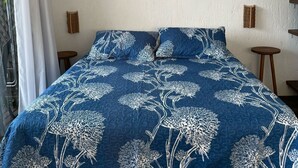 A bedroom with a blue floral bedspread, a white brick-patterned headboard, a wall-mounted air conditioner, and a decorative wall hanging. - Altos da Lagoa-praia do Rosa,ibiraquera Studio 1 Para Casais Calmo e Tranquilo (imbituba)