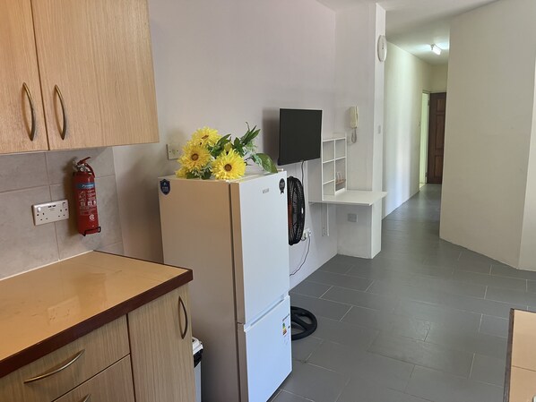 A kitchen with a white refrigerator, a fire extinguisher, and a wall-mounted fan. - Sunset Plaza Apartments 4 (Victoria)
