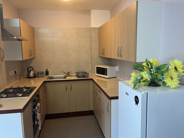 A compact kitchen with wooden cabinets, a microwave, a kettle, and a refrigerator.