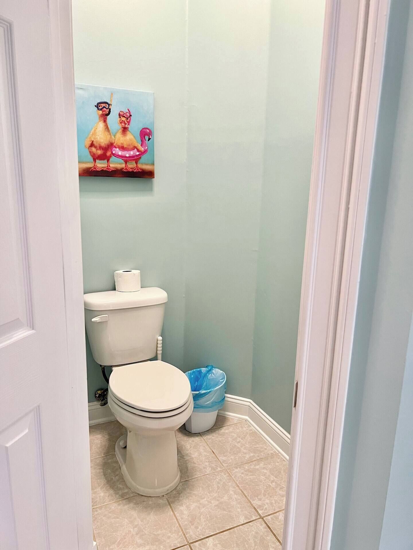 A small bathroom with a toilet, a blue trash bin, and a painting of chickens on the wall.