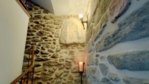 A staircase with stone walls and a metal railing, leading to a room with a stone wall and a lit candle on a wall-mounted holder.