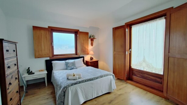 A bedroom with a bed, wooden wardrobe, a nightstand, a dresser, a window, and a view of the sea.