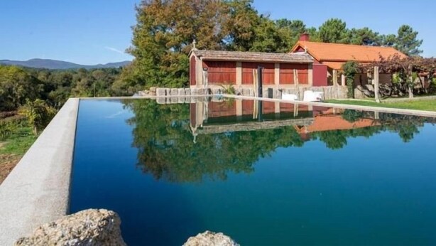 A swimming pool with a clear reflection, a red building with a tiled roof, and surrounding greenery. - Quinta sw Vila Nova | Piscina | Agrotourism | Rural Environment (Monção)