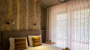 A bedroom with a stone wall, a bed with striped pillows, a nightstand, and a window with sheer curtains. - Quinta sw Vila Nova | Piscina | Agrotourism | Rural Environment (Monção)