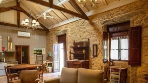 A rustic interior with a stone wall, wooden beams, a sofa, a dining table, and a kitchen area. - Quinta Vila Nova | Apartamento das Camélias | Agrotourism | Rural Environment (Monção)