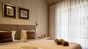 A hotel room with two beds, a headboard, a nightstand, and a window with sheer curtains.