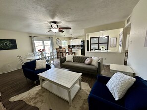 Ein Wohnzimmer mit einer Couch, Sesseln, einem Couchtisch, einem Deckenventilator und einer Küche im Hintergrund.