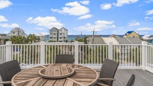 A wooden deck with a round table and wicker chairs, overlooking a beach with buildings in the background. - 4806 Hispaniola St. 3 Kings - 4 Twins - Private Heated Pool! 2 Minute Walk to the Sand! (Panama City Beach)
