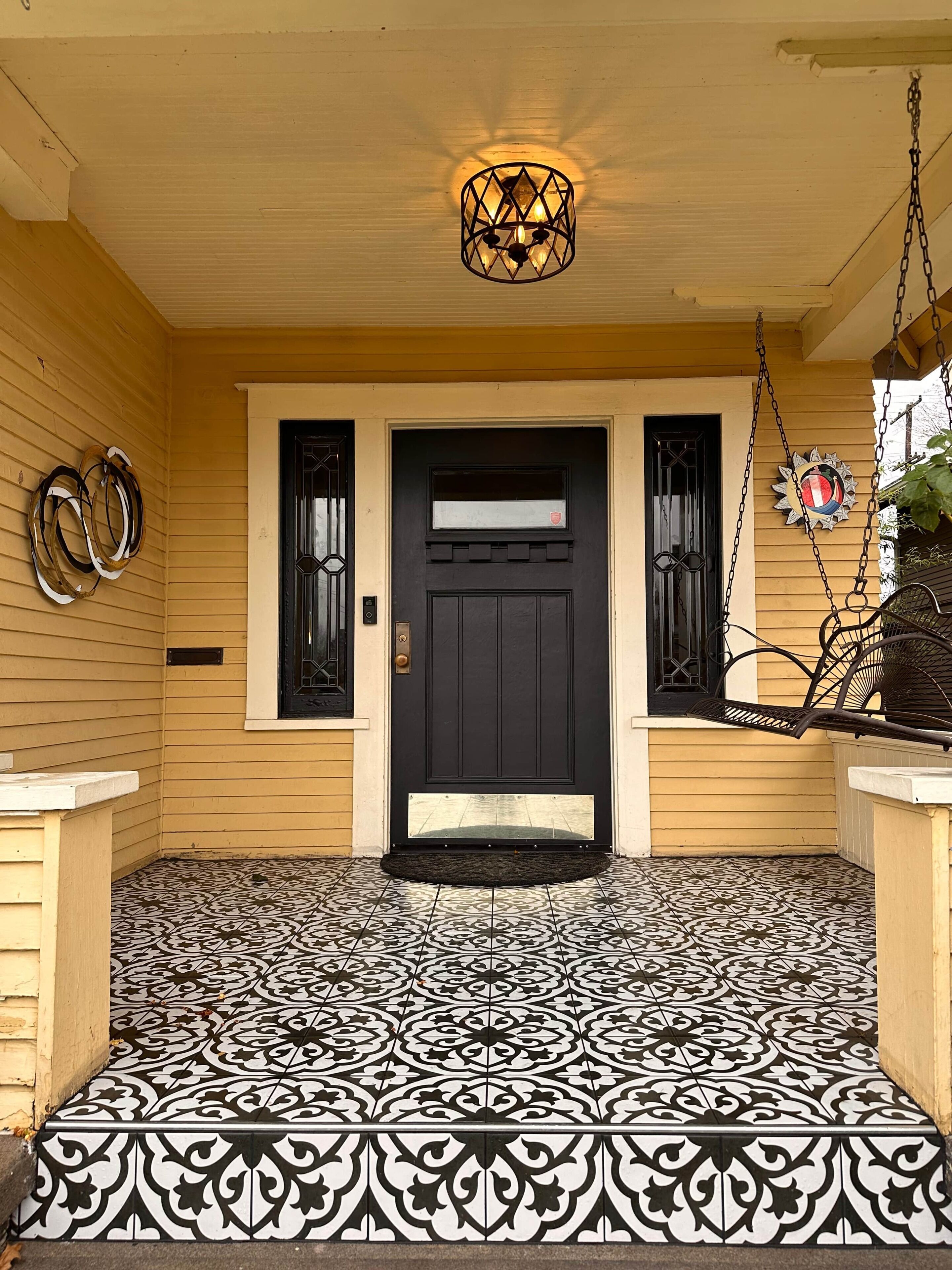 Un porche à l’avant de la maison, avec une porte noire, un luminaire suspendu et un sol carrelé à motifs.