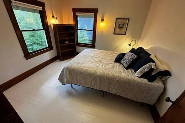Une chambre à coucher avec un lit, deux fenêtres, une table de chevet et un luminaire fixé au mur.