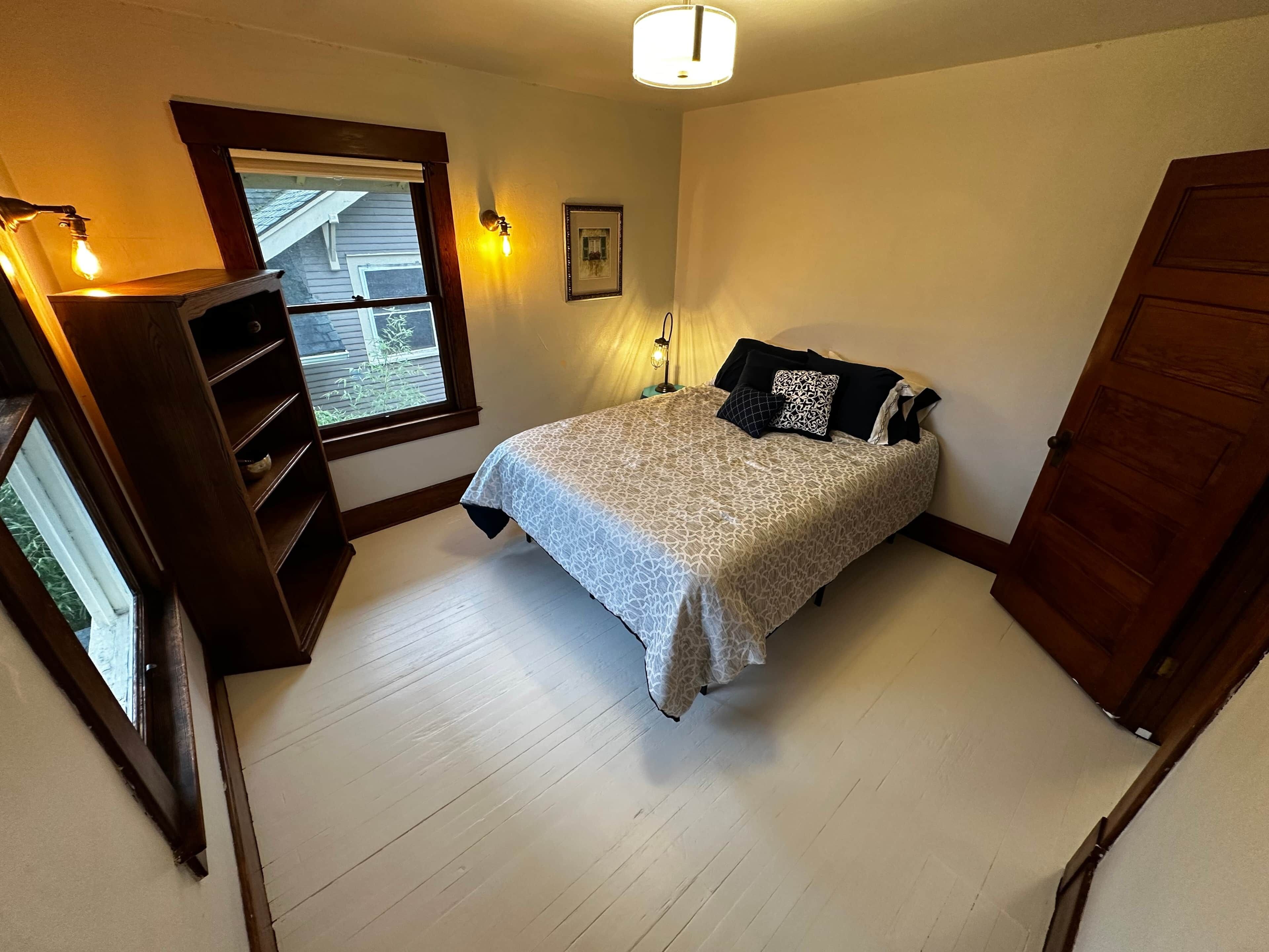 Un dormitorio con una cama, una mesita de noche, una ventana, una puerta y una lámpara de pared.