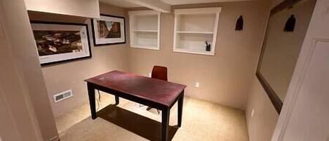 A small room with a dark wooden table and a single red chair. There are two white shelves on the wall, and a framed picture is visible.