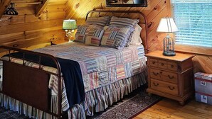 A cozy attic bedroom with a sloped ceiling, a bed with a patterned bedspread, a nightstand with a lamp, and a window with blinds. - Cardinal Nest -Mountain views, babbling stream, fields to play in, privacy! (Blairsville)