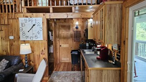 A cozy cabin interior with a wooden staircase, a dining area, a kitchen with wooden cabinets, and a large clock on the wall. - Cardinal Nest -Mountain views, babbling stream, fields to play in, privacy! (Blairsville)