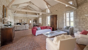 A spacious living area with a stone wall, wooden beams, and a brick floor. There is a sofa, a coffee table, and a fireplace.