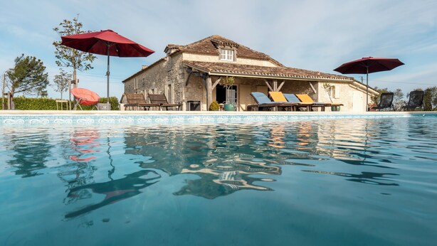 A pool area with lounge chairs and umbrellas, a stone building with a tiled roof, and a clear blue pool.