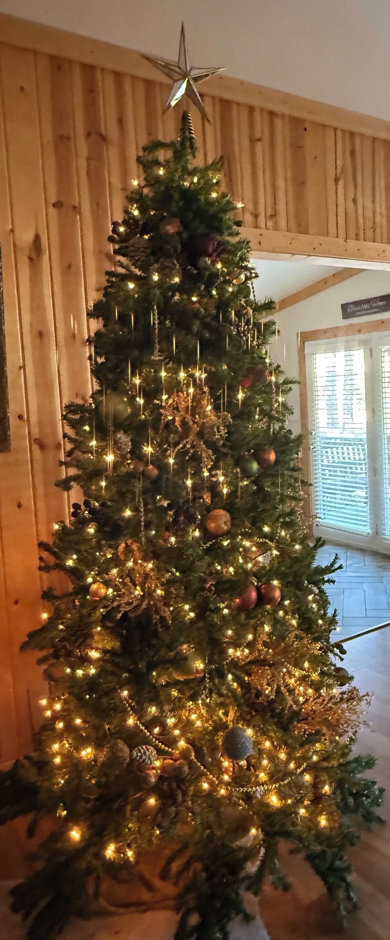 Un sapin de Noël décoré de guirlandes lumineuses et d’ornements, dans une pièce aux murs en bois et dotée d’une fenêtre.