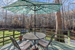 A round table with four chairs and a large umbrella on a deck with a green railing, surrounded by trees.