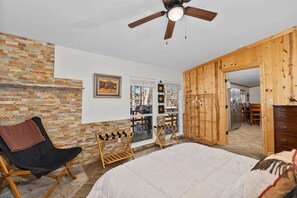 A bedroom with a large bed, a ceiling fan, a brick wall, wooden furniture, and a view of the dining area.