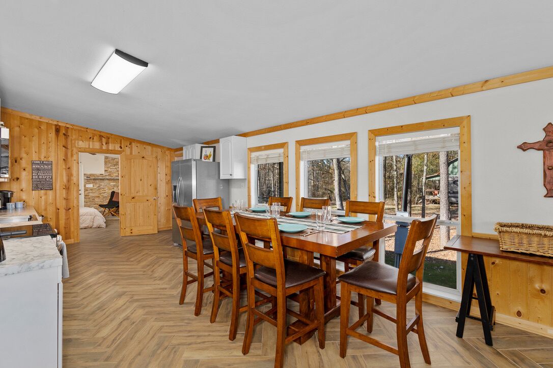 Une salle à manger aux murs lambrissés, avec une grande table dressée pour six personnes, flanquée de fenêtres équipées de stores, d’un réfrigérateur et d’un comptoir de cuisine visible au fond.