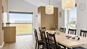 Uma área de jantar moderna com uma mesa grande, cadeiras, luminárias pendentes e vista através de uma porta de vidro.