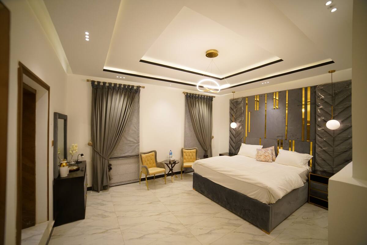 A modern bedroom with a large bed, two chairs, a small table, and a mirror.