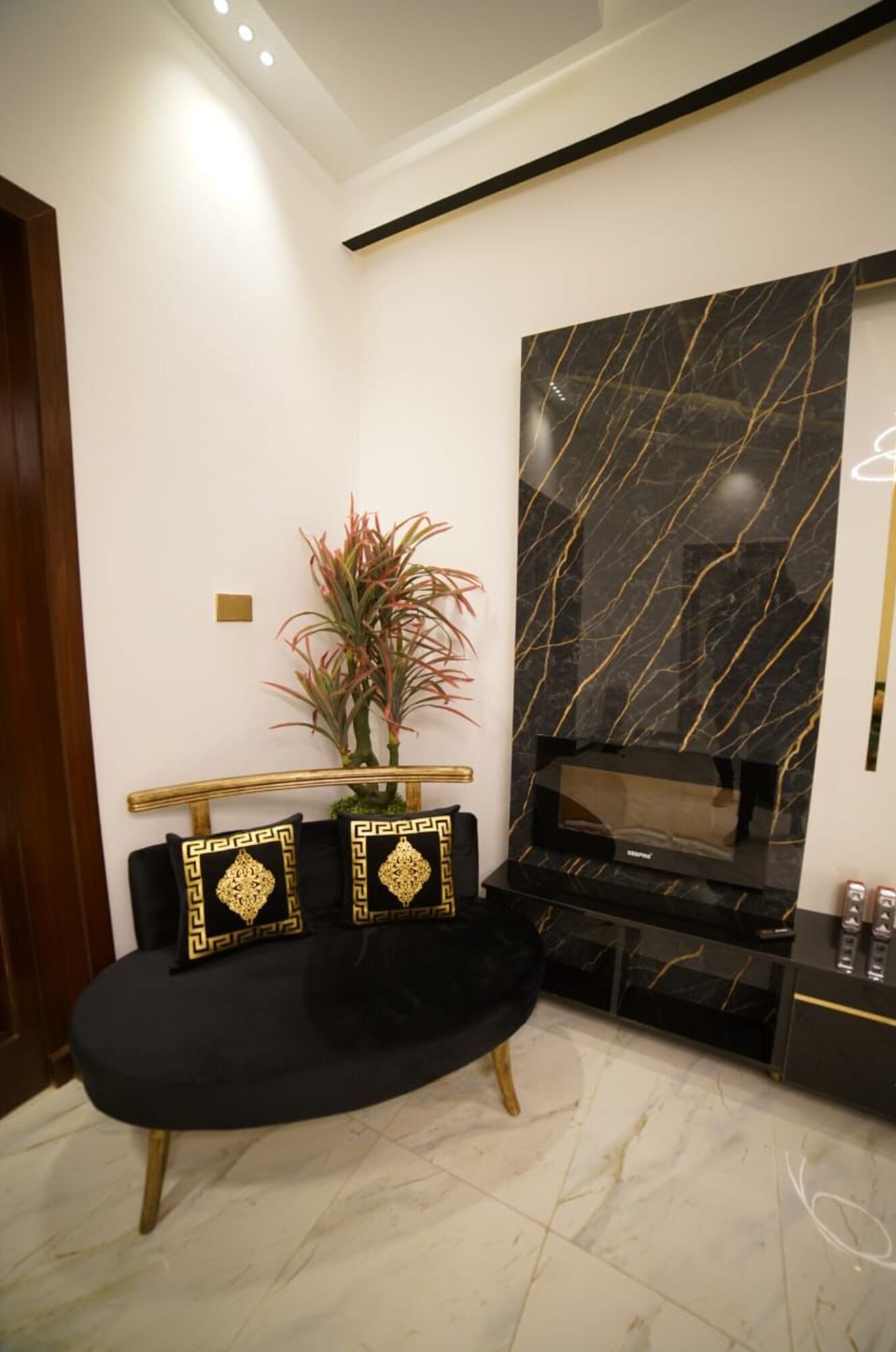 A modern living room with a black sofa, gold decorative pillows, a marble-patterned wall, and a small plant.