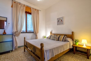 A bedroom with a wooden bed, a bedside table with a lamp, a window with curtains, a small plant, and a framed picture on the wall.